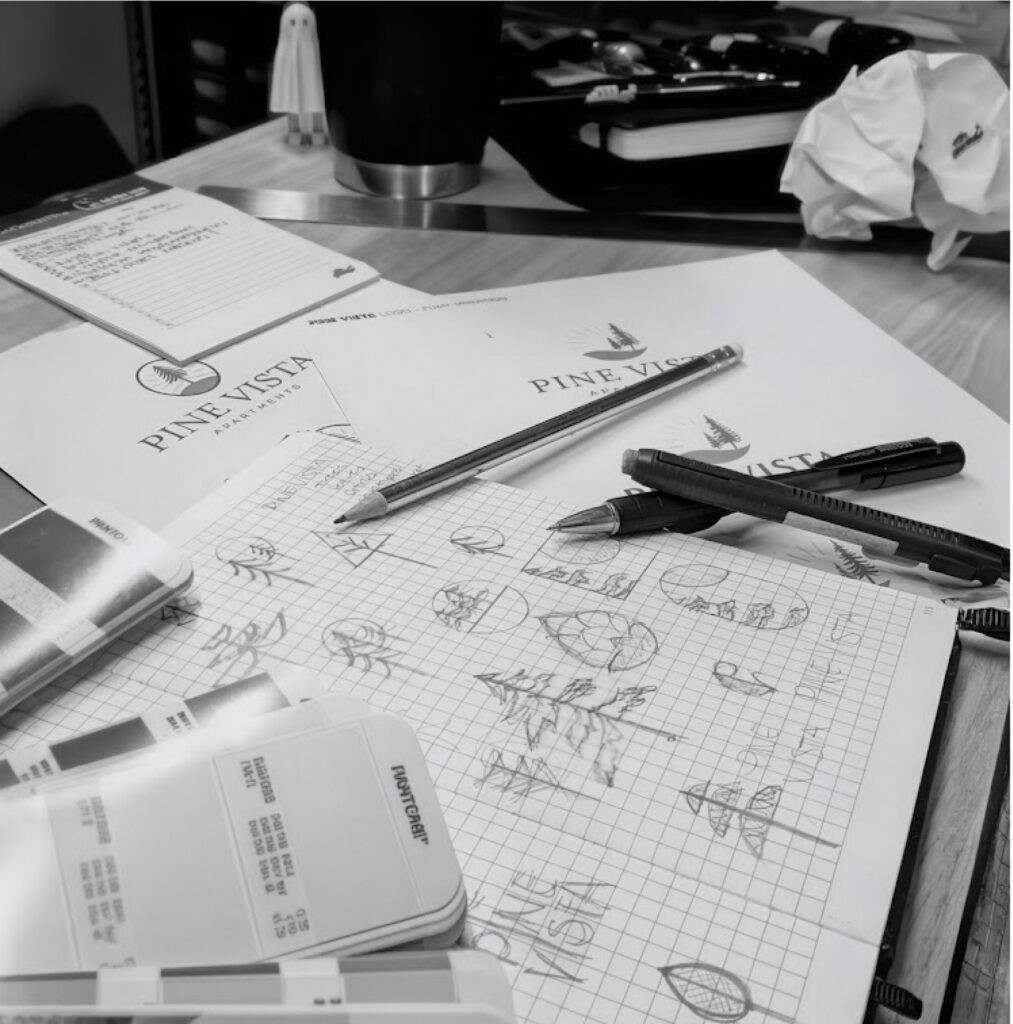 flat lay of design materials on a desk — logo sketches, pantone color swatches, pencils, erasers, coffee mug, printed logo mockups, ruler